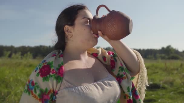 Söt överviktiga kvinnan njuter av att dricka färsk mjölk från jord kannan på den gröna sommaren fältet. Vackert landskap. Folklore, traditioner koncept. Verklig lantlig kvinna. — Stockvideo