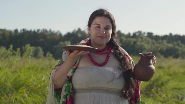 Belle femme en surpoids reniflant des crêpes et buvant du lait frais du pot de terre sur le champ vert d'été. Beau paysage. Style de vie rural. Femme rurale réelle . — Video