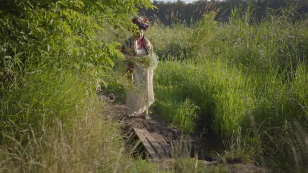 Mooi overgewicht vrouw met een krans op haar hoofd en bloemen in handen gaan door de kleine oude brug van de planken. Landelijke levensstijl. Verband met de natuur, het plattelandsleven. — Stockvideo