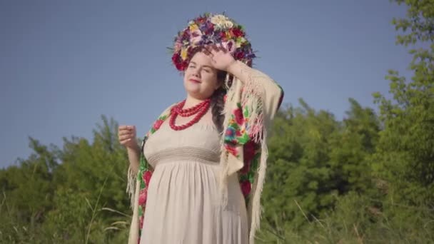 Porträt einer süßen dicken Frau mit einem Kranz auf dem Kopf, die im Sonnenlicht auf der grünen Sommerwiese lächelt. Verbindung mit der Natur. Echte Landfrau. — Stockvideo