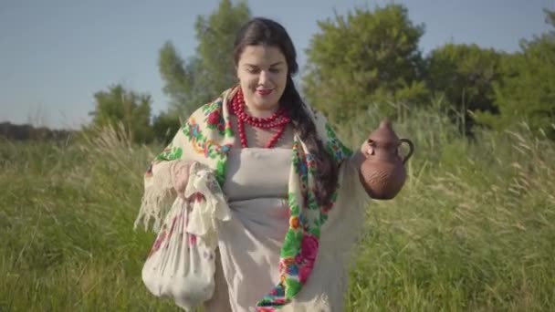 Belle femme en surpoids marchant dans l'herbe sauvage haute avec le pot de terre dans le champ vert d'été. Beau paysage. Concept de traditions, lien avec la nature. Style de vie rural — Video