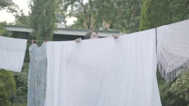 Cute mature woman with long hair hanging white clothes on a clothesline looking at camera outdoors. Washday. Lady doing laundry. Concept of sustainability, nature and purity and deep clean after — Stock Video