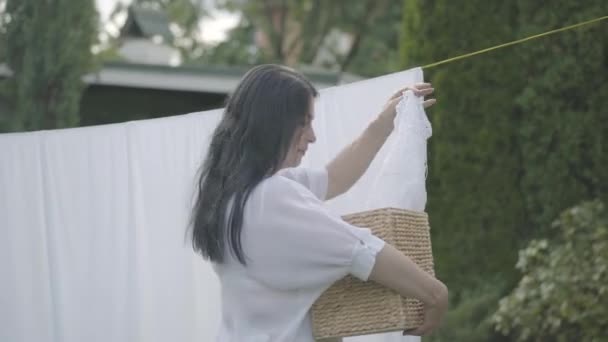 Mature housewife woman is hanging just washed clean laundry on clothesline outside her house in a sunny day. Concept of sustainability, nature and purity and deep clean after washing. — Stock Video