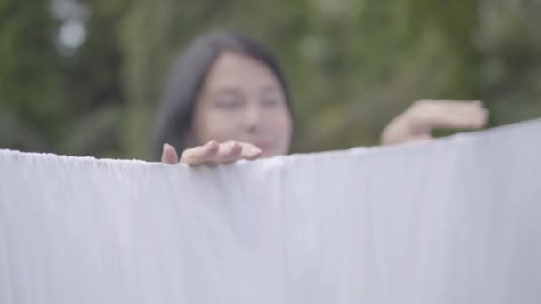 Portret schattig volwassen vrouw met lange haren opknoping witte kleren op een waslijn kijken naar camera buitenshuis. Wasdag. Dame doen Wasserij. Concept duurzaamheid. — Stockvideo