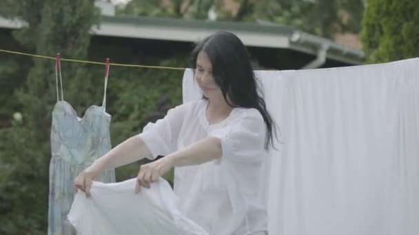 Jolie femme âgée aux cheveux longs cheveux noirs suspendus vêtements blancs sur une corde à linge à l'extérieur. Le jour de toilette. Femme au foyer positive faisant la lessive — Video