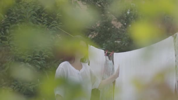 Niedlichen reifen Frau mit langen Haaren hängen weiße Kleidung auf einer Wäscheleine im Freien. Eine erwachsene Frau bindet sich ein Tuch an den Kopf und blickt in die Kamera. Waschtag. Dame beim Wäschewaschen. Konzept der Nachhaltigkeit — Stockvideo