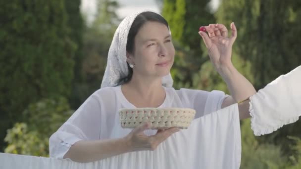 Hübsche Frau mit weißem Schal auf dem Kopf, die Kirschen isst und über die Wäscheleine im Freien in die Kamera lächelt. Waschtag. positive Hausfrau beim Wäschewaschen — Stockvideo