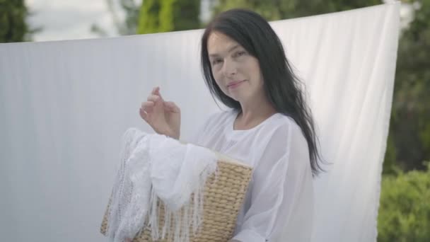 Adorable mature woman with long black hair holding wicker basket in hands while hanging white clothes on a clothesline outdoors and looking at camera. Washday. Senior woman doing laundry. — Stock Video