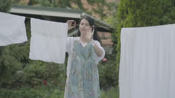 Attraktive reife Frau mit langen Haaren, die ihre Kleidung an einer Wäscheleine im Freien aufhängt, dann das Kleid anprobiert und lächelnd in die Kamera schaut. Waschtag. Dame beim Wäschewaschen — Stockvideo