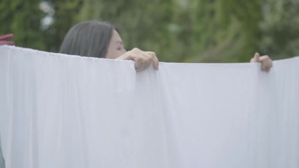 Όμορφη ώριμη γυναίκα με μακριά μαλλιά κρέμονται λευκά ρούχα σε μια γραμμή άπλων κοιτάζοντας την κάμερα σε εξωτερικούς χώρους. Μπουγάδας. Η κυρία κάνει μπουγάδα. Έννοια της αειφορίας. — Αρχείο Βίντεο