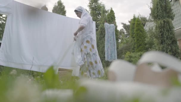 Mulher dona de casa madura pendurado apenas lavou a roupa limpa no varal fora de sua casa em um dia ensolarado. Conceito de sustentabilidade, natureza e pureza e limpeza profunda após lavagem . — Vídeo de Stock