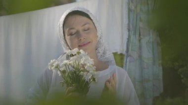 Kafasında beyaz şal olan sevimli kadın, çamaşır ipinin yanındaki kameraya bakarak papatyaları kokluyor. Yıkanıyor. Pozitif kaygısız ev hanımı çamaşır yapıyor