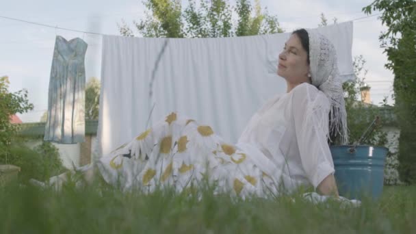 Mulher sênior atraente com um xale branco na cabeça descansando no jardim sentado na grama na frente do varal. Lavabo. Dona de casa positiva descansando depois de lavar a roupa. Vista inferior — Vídeo de Stock