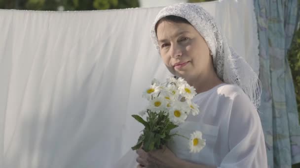 Portret starszy kobieta z białym szal na głowie wąchania stokrotki patrząc na kamerę w pobliżu bielizny na zewnątrz. Na dzień. Pozytywna beztroski gospodyni robi pranie — Wideo stockowe
