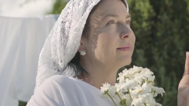 彼女の頭の上に白いショールを持つかわいい成熟した女性は、屋外の衣服ラインでデイジーの花びらを引き裂きます。ウォッシュデイ洗濯をしているポジティブな気楽な主婦。木のブランチの後ろから撮影 — ストック動画