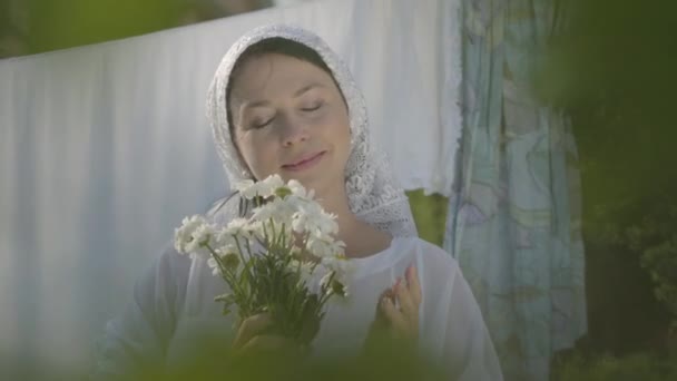 Adorable woman with white shawl on her head sniffing daisies looking at camera near the clothesline outdoors. Washday. Positive carefree housewife doing laundry — Stock Video
