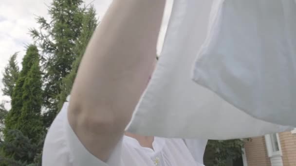 Porträt einer attraktiven Seniorin mit weißem Tuch auf dem Kopf, die Bettwäsche aus dem Seil zieht. Waschtag. positive Hausfrau beim Wäschewaschen. von unten — Stockvideo