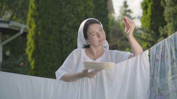 Mujer bonita con chal blanco en la cabeza comiendo cerezas mirando a la cámara sonriendo sobre el tendedero al aire libre. Lavado. Ama de casa positiva lavando ropa. Movimiento lento . — Vídeo de stock