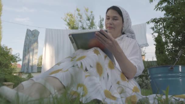 Atractiva mujer mayor con un chal blanco en la cabeza descansando en el jardín sentado en la hierba frente a la revista de lectura de tendedero. Ama de casa positiva descansando después de lavar la ropa. Vista inferior — Vídeo de stock