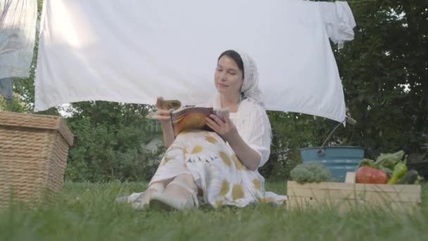 Adorabile donna con uno scialle bianco sulla testa che riposa in giardino seduta sull'erba davanti alla rivista di lettura clothesline. Casalinga positiva a riposo dopo aver fatto il bucato. Vista dal basso — Video Stock