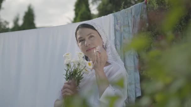 그녀의 머리에 흰색 목도리를 가진 예쁜 선임 여성은 야외에서 옷라인에서 데이지 꽃잎을 찢습니다. 워시데이. 긍정적 인 평온한 주부 세탁을하고. 나무 브런치 뒤에서 촬영 — 비디오