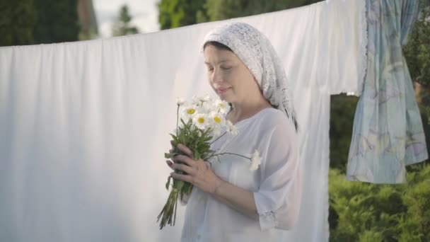 Cute mature woman with white shawl on her head tears off daisy petals at the clothesline outdoors. Washday. Positive carefree housewife doing laundry. Shooting from behind tree brunch. Slow motion. — Stock Video