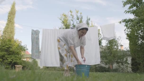 Attractive senior woman with a white shawl on her head hanging bed linen on the rope in the garden close-up. Washday. Positive housewife doing laundry. — Stock Video