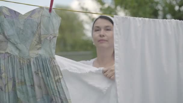 Porträtt trött söt mogen kvinna hängande vita kläder på en klädstreck tittar på kamera utomhus. Gick. Lady gör tvätt. Hållbarhets begrepp. — Stockvideo