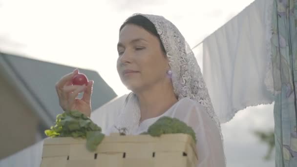 Aantrekkelijke Senior vrouw met een witte sjaal op haar hoofd houden de mand met groenten snuiven Cherry tomaat in de buurt van de waslijn buitenshuis. Positieve huisvrouw doet huiswerk — Stockvideo