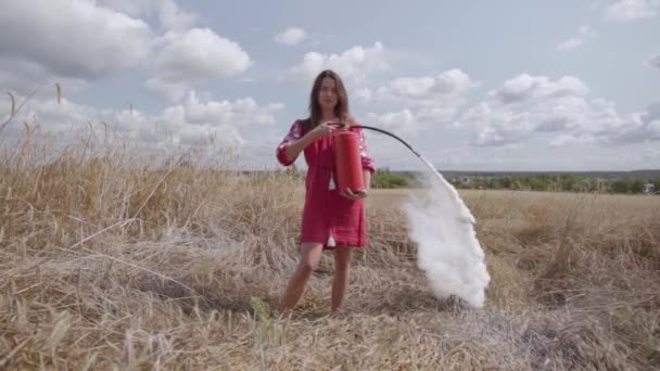 Atraktivní žena s dlouhými vlasy, která používá hasicí přístroj na pšeničné pole. Souvislost s přírodou, přírodní krásou. Čas sklizně. Nebezpečí požáru, koncepce protipožární prevence. Zpomaleně. — Stock video
