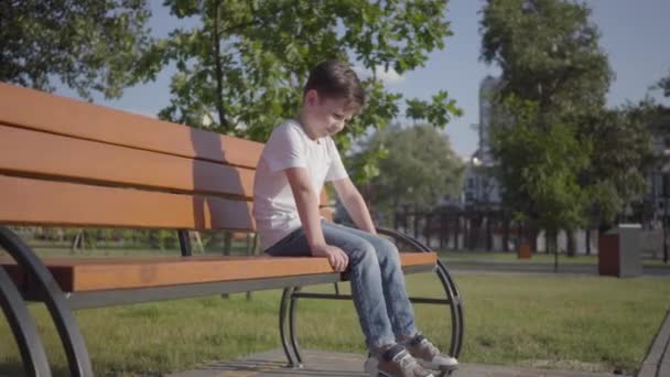 Сумний самотній хлопчик сидить на лавці в парку. Мила дитина проводить час один на відкритому повітрі. Літній відпочинок — стокове відео
