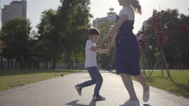 Joyful older sister spinning around with younger brother holding hands in the summer park. Leisure outdoors. Friendly relations between siblings. Carefree kids having fun together — Stock Video