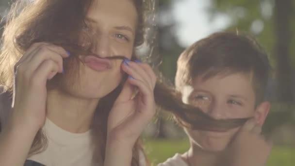 Retrato da irmã mais velha passar tempo com o irmão mais novo ao ar livre. O menino e a menina que fazem bigodes falsos com o cabelo da menina no parque. Lazer. Os irmãos se divertem juntos — Vídeo de Stock