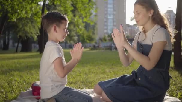 Äldre syster spenderar tid med yngre bror utomhus. Pojken och flickan som spelar Pat-a-Cake sitter på filten i parken. Sommar fritid. — Stockvideo