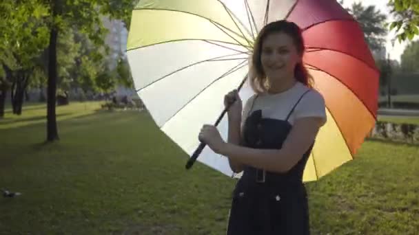 Jeune fille mignonne avec parapluie multicolore tournant et regardant la caméra souriant dans le parc. Loisirs d'été. Jeune femme s'amuser à l'extérieur . — Video