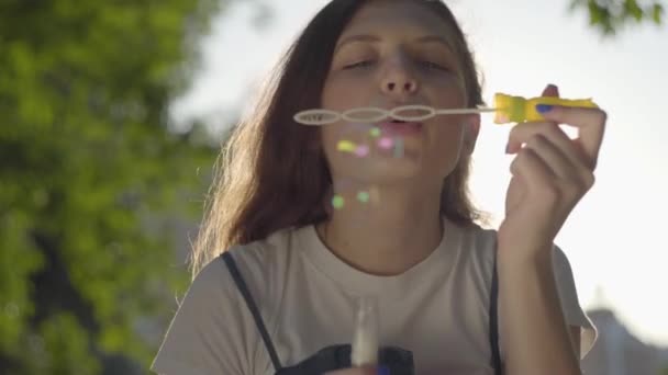 Portrait of the teenage girl in the park blowing soap bubbles at the camera. Cute young woman spending time alone outdoors. Summertime leisure — Stock Video