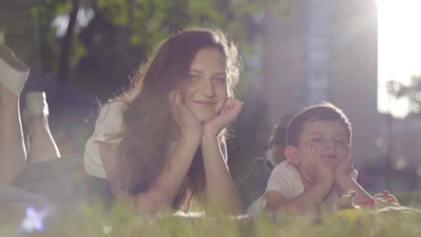 Портрет старшої сестри та молодшого брата на відкритому повітрі. Хлопчик і дівчинка лежать на траві разом у парку, дивлячись на камеру. Літнє дозвілля. Прекрасна літня погода, сонячне світло . — стокове відео