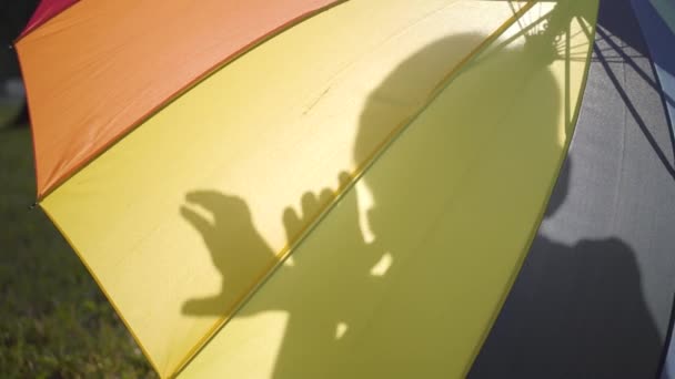 Різнокольорова парасолька лежить в парку на траві. Силует маленької безтурботної дитини, що сидить за парасолькою і показує фігури руками. Літнє дозвілля — стокове відео