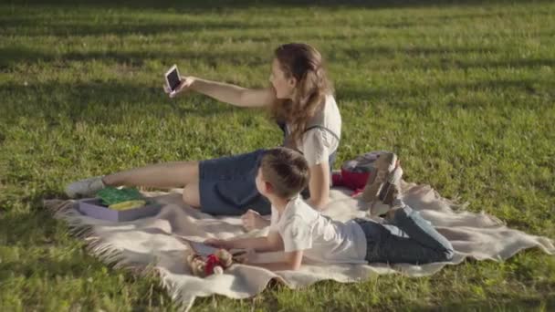 Die ältere Schwester verbringt Zeit mit dem jüngeren Bruder im Freien. der Junge beim Lesen des Buches und das Mädchen beim Selfie im Park. Sommerfreizeit. Spielzeugsucht — Stockvideo