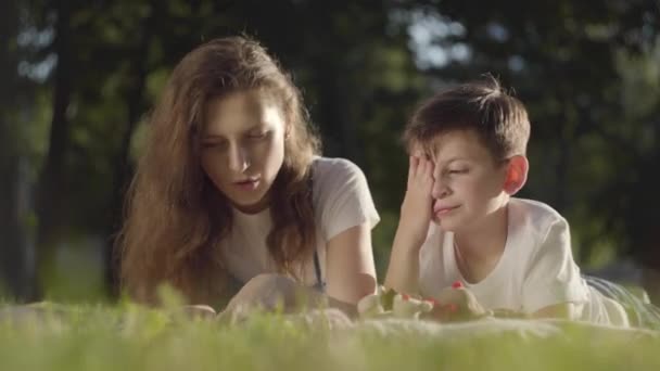 Portrait d'une sœur aînée passant du temps avec son frère cadet à lire un livre ou un magazine à l'extérieur. Le garçon et la fille couchés sur l'herbe ensemble dans le parc. Loisirs d'été — Video