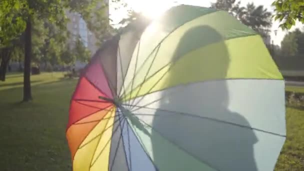 Привлекательная молодая девушка с разноцветным зонтиком, поворачивающимся и смотрящим на улыбающуюся в парке камеру. Летний отдых. Молодая леди веселится на открытом воздухе — стоковое видео