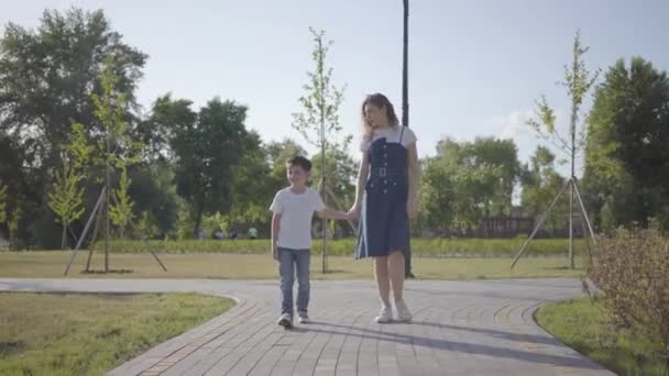 Starsza siostra spaceru z młodszym bratem trzymając ręce w parku letnim. Niegrzeczny chłopiec ucieka, Dziewczyna biegnie po nim. Wypoczynek na świeżym powietrzu. Przyjazne stosunki między rodzeństwem — Wideo stockowe