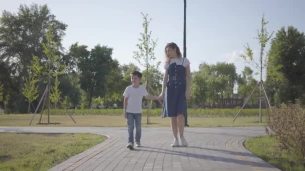 Die ältere Schwester geht mit dem jüngeren Bruder Händchen haltend durch den Sommerpark. Der freche Junge rennt weg, das Mädchen rennt ihm hinterher. Freizeit im Freien. Freundschaftliche Beziehungen zwischen Geschwistern — Stockvideo