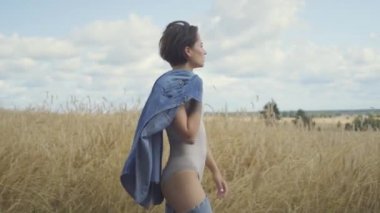 Çekici bir kadının omuz buğday alanında yürürken kısa saç tutan kot ceket ile bodysuit giyen yan görünümü. Kendine güvenen kaygısız kız açık havada. Gerçek insanlar serisi.