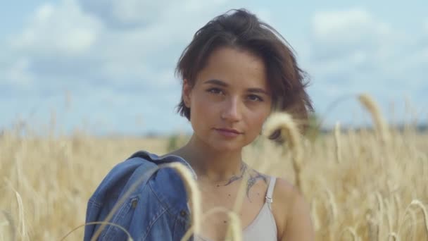 カメラをクローズアップして見て小麦畑に立つジーンズジャケットを持つ短い髪の魅力的な女性の肖像画。自然とのつながり、自然の美しさ — ストック動画