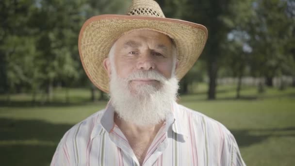 Portrait old grey-haired bearded man in straw hat looking at camera smiling standing in the park. Leisure outdoors. Mature man resting in the summer garden. Healthy cheerful senior retired man — Stock Video