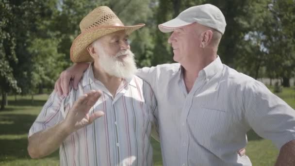 Två positiva gamla män som tittar i kameran viftar händerna i parken. Fritid utomhus. Äldre människor vilar i sommarträdgården. Senior manliga vänner tillsammans. Friska glada Senior pensionerade män. — Stockvideo