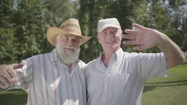 Two handsome old men looking in the camera waving hands in the park. Leisure outdoors. Mature people resting in the summer garden. Senior male friends together. Healthy cheerful senior retired men. — Stock Video