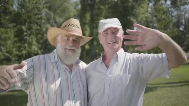 Twee knappe oude mannen op zoek in de camera zwaaiende handen in het Park. Vrije tijd buitenshuis. Volwassen mensen rusten in de zomertuin. Senior mannelijke vrienden samen. Gezonde vrolijke Senior gepensioneerde mannen. — Stockvideo