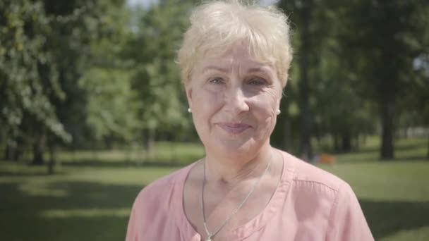 Ritratto vecchia donna dai capelli grigi guardando la macchina fotografica in piedi nel parco estivo. Tempo libero all'aperto. Maturo signora caucasica a riposo in giardino. sana allegra anziana pensionato signora — Video Stock